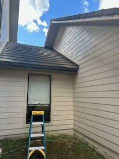 A yellow and blue step ladder stands against the light-colored siding of a house below a tile roof and a window.