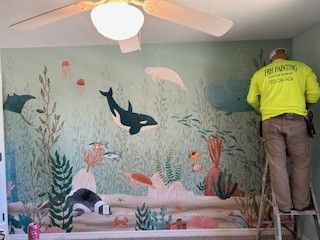 A person stands on a ladder, painting a vibrant underwater mural featuring a whale, stingray, and colorful sea life.