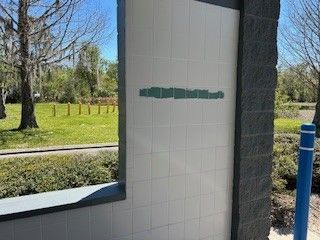 A light-colored tiled wall with a decorative line of green tiles, set outdoors near trees and a blue bollard.