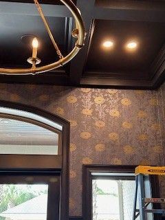 A dark coffered ceiling with a gold chandelier and recessed lighting above patterned wallpaper and two dark-framed windows.