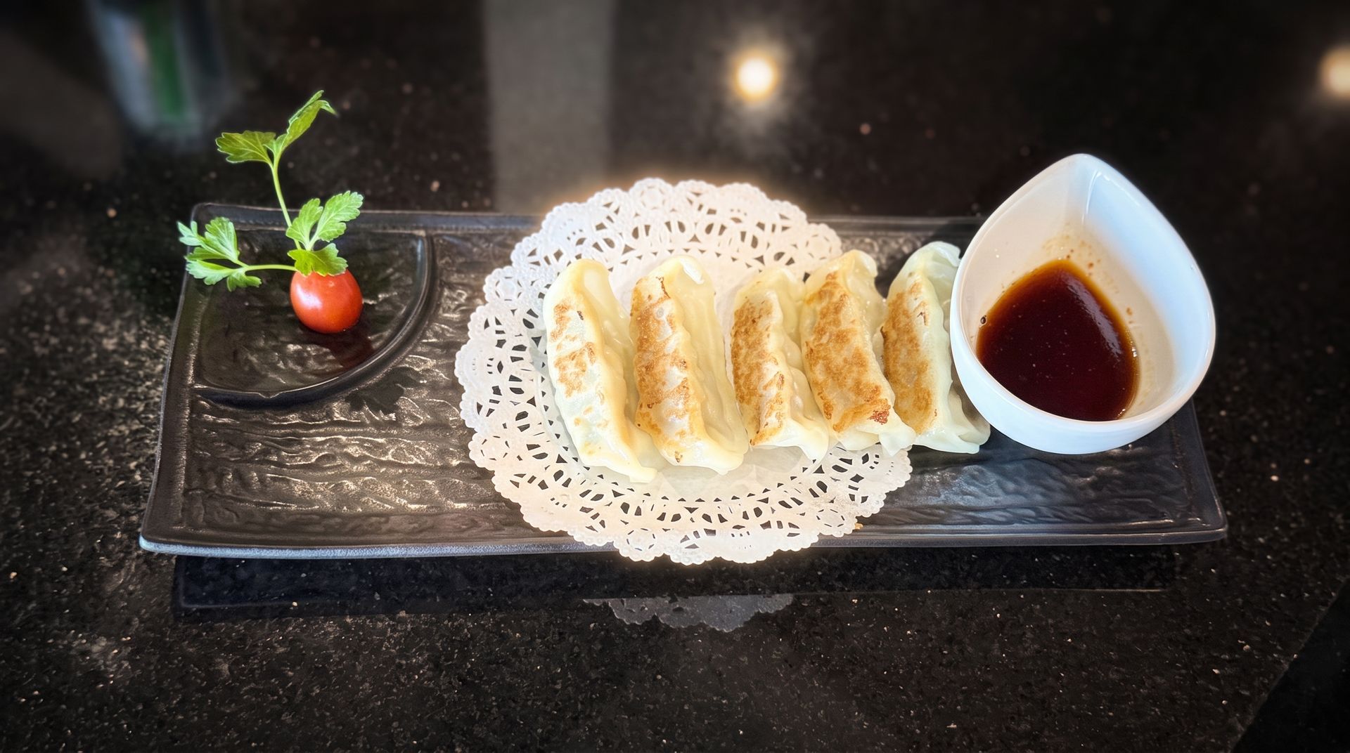 Fried spring rolls on a doily with dipping sauce and a cherry tomato garnish on a black tray.