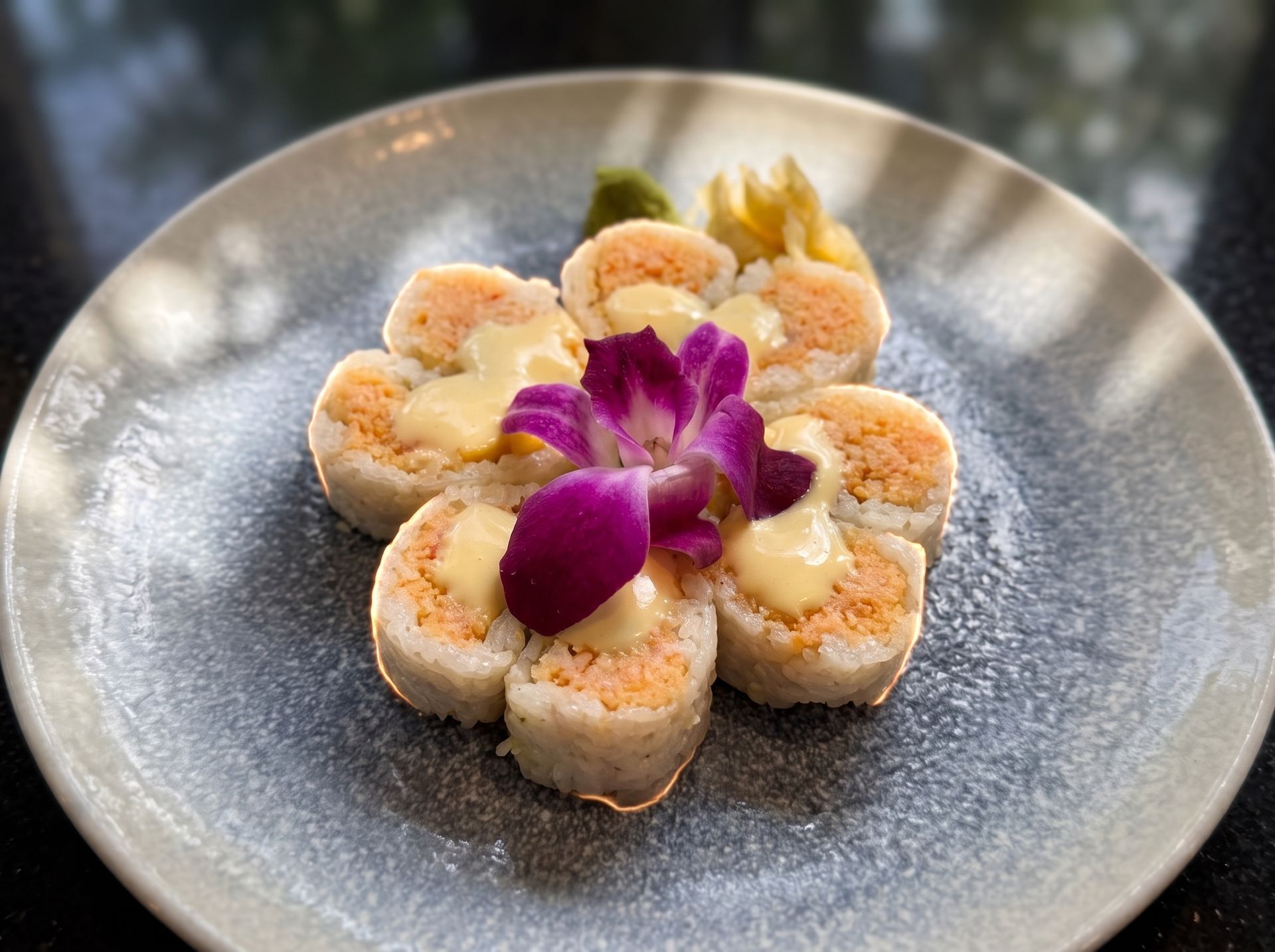 Sushi rolls arranged on a gray plate, topped with purple orchid flowers and sesame seeds.