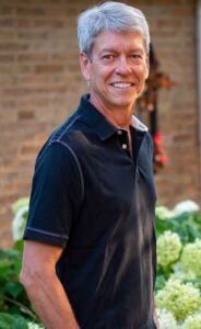 A smiling person wearing a dark polo shirt stands in front of a brick wall and green flowering plants.