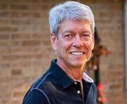 A person with grey hair smiles, wearing a dark polo shirt while standing outdoors near a brick wall and green foliage.