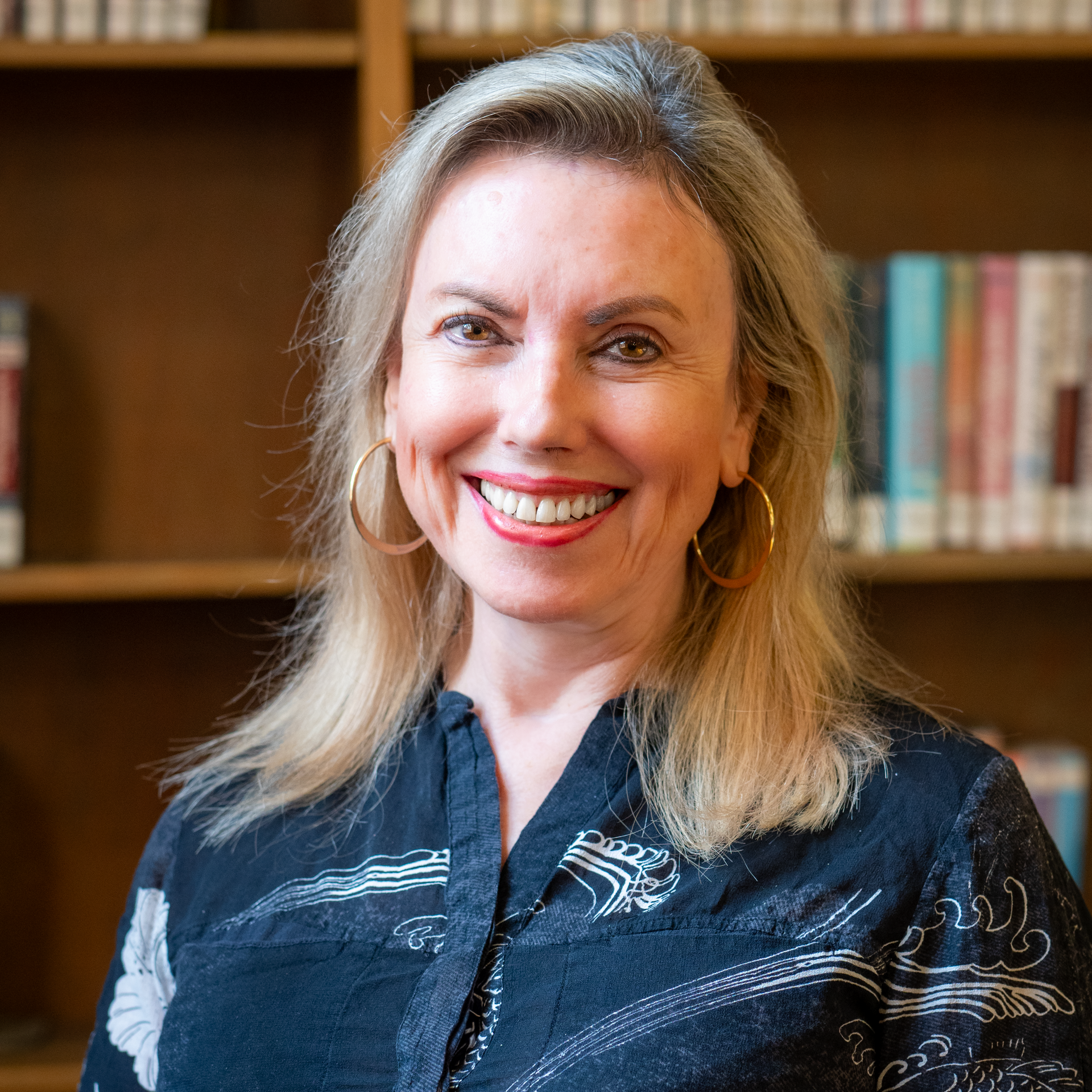 A smiling person with blonde hair and hoop earrings, wearing a dark patterned shirt, standing in front of bookshelves.