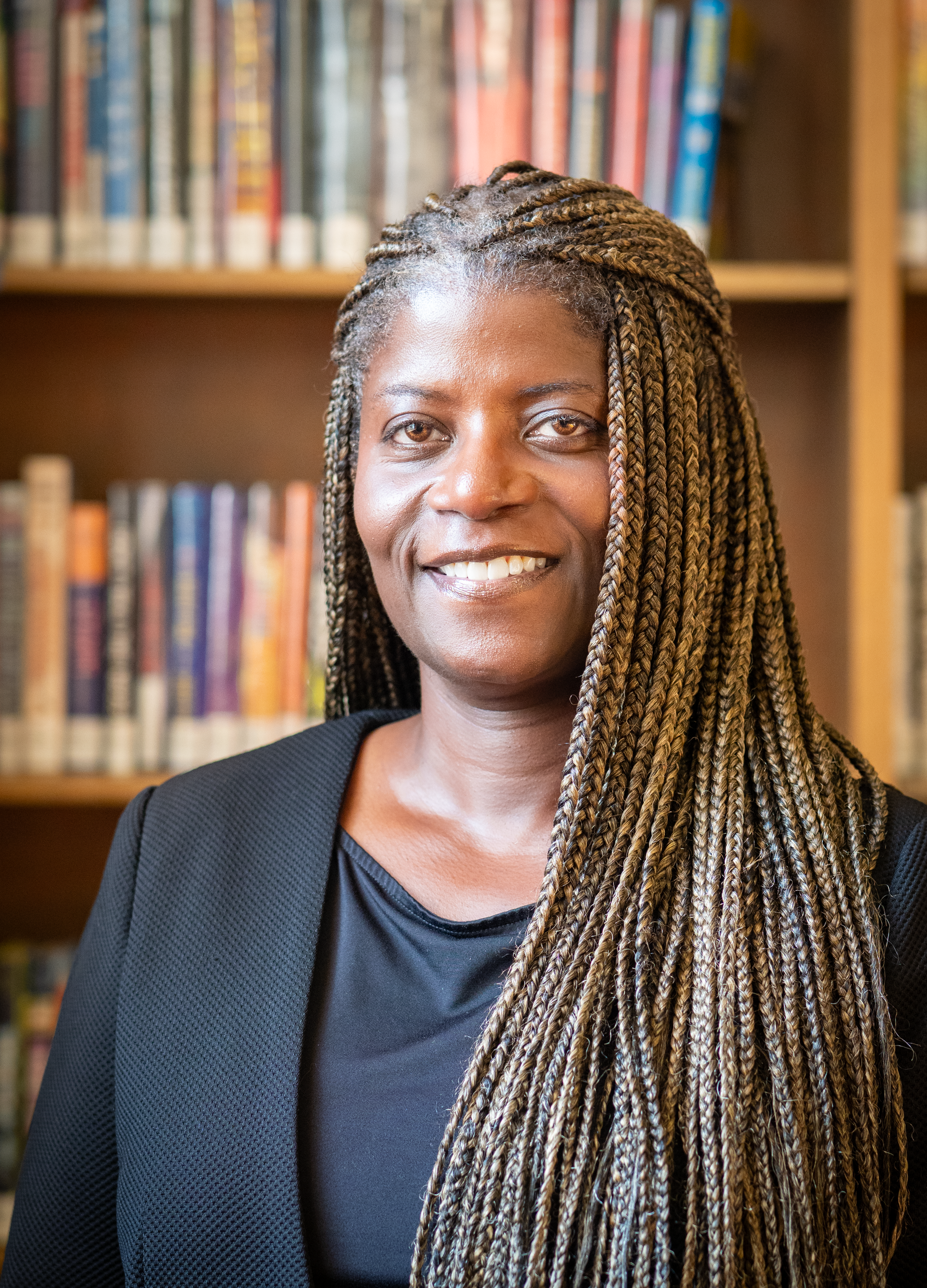 A smiling person with long braided hair wearing a black blazer, posed in front of a bookshelf filled with books.