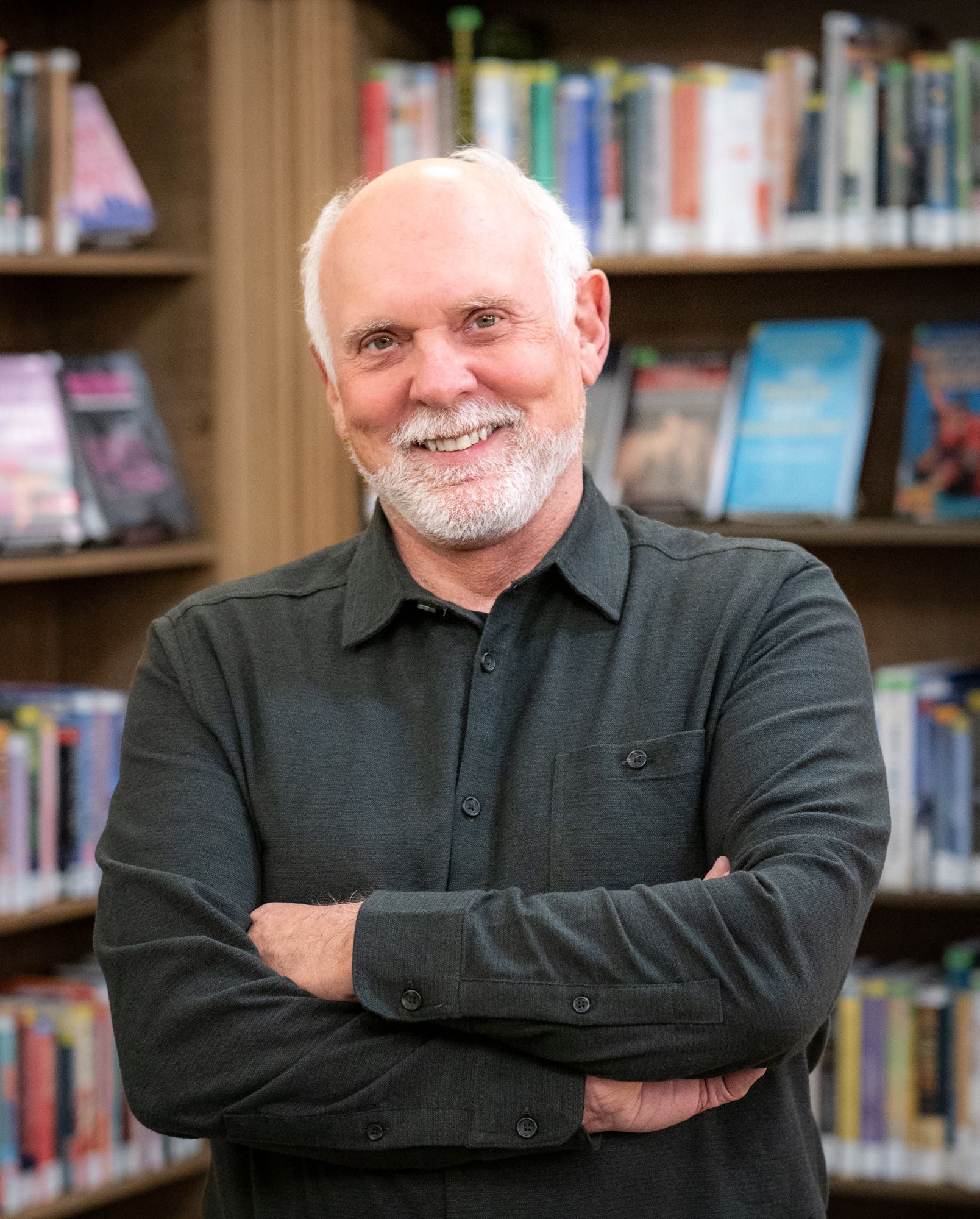 A person with a beard and gray hair wearing a dark button-down shirt, standing with arms crossed in a library.
