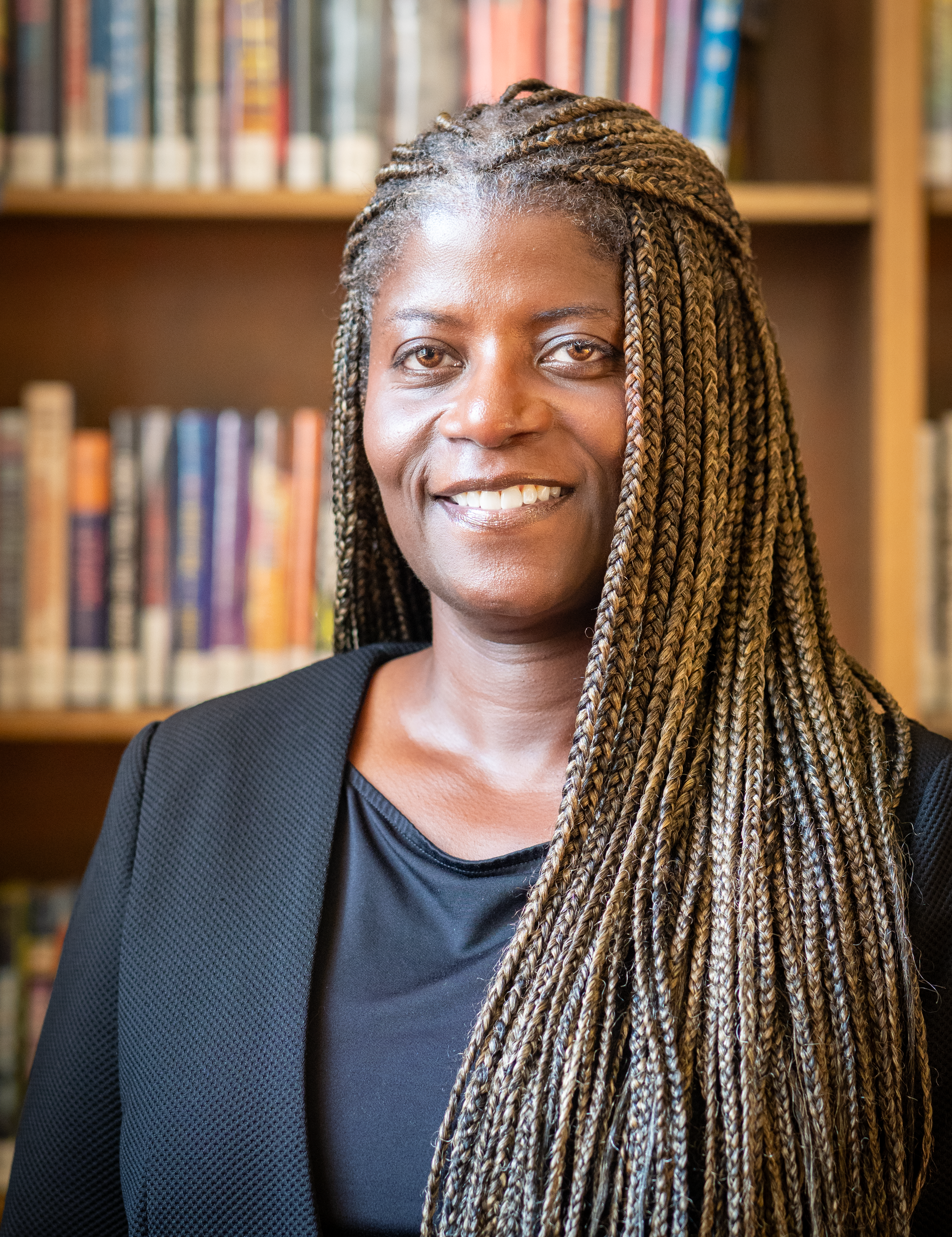 A smiling individual with long braided hair wearing a dark blazer, positioned in front of a library bookshelf.