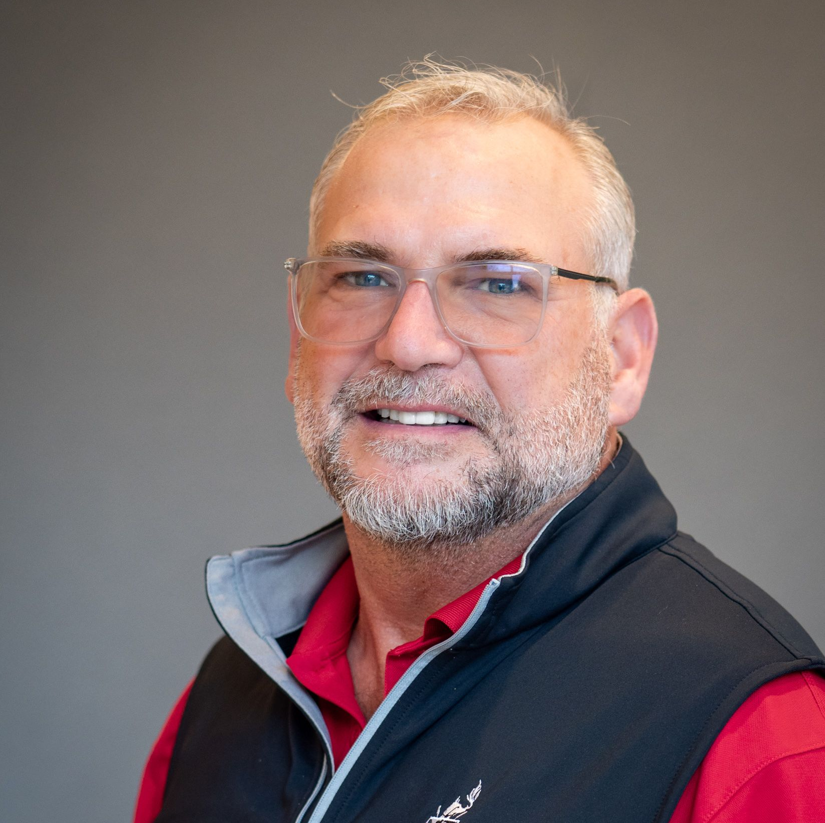 A smiling person with glasses, a gray beard, and a red shirt under a dark blue quarter-zip vest against a gray background.