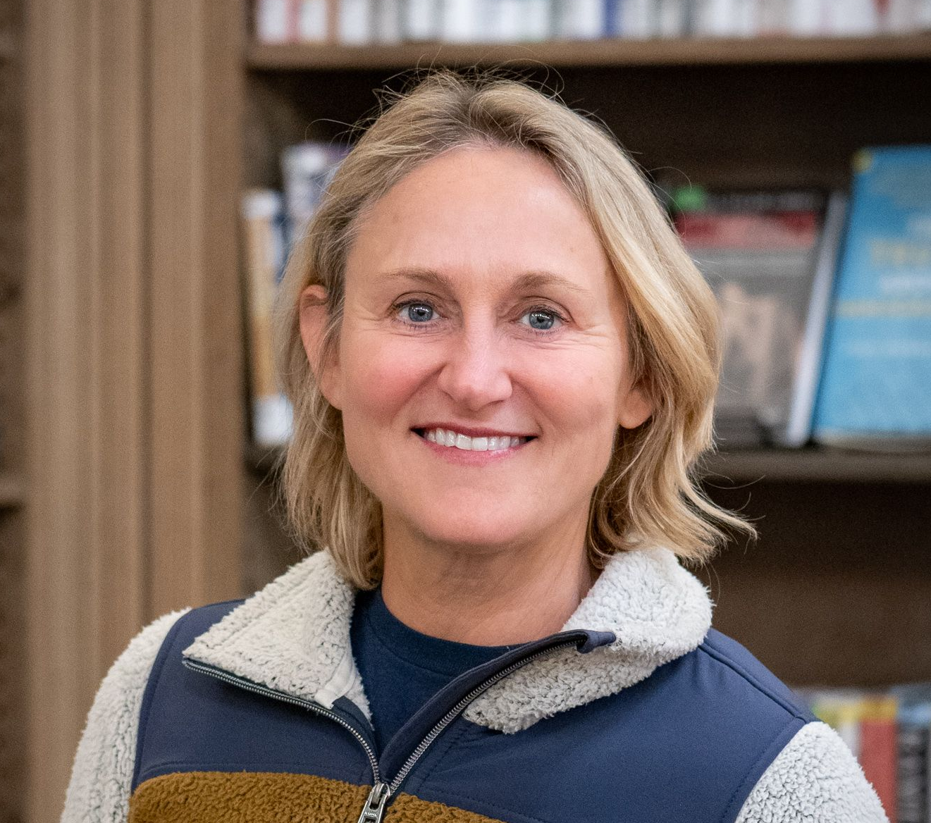 Smiling person wearing a mustard and navy fleece jacket, standing in front of a bookshelf filled with books.