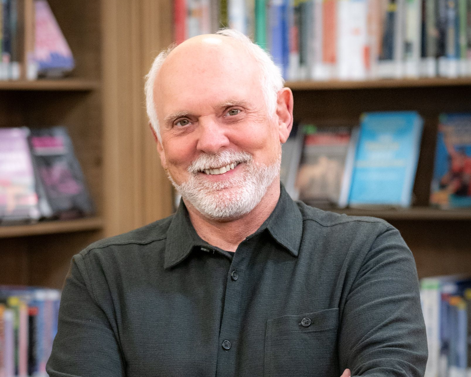 A person with a white beard and hair smiles while standing with arms crossed in front of a bookshelf.