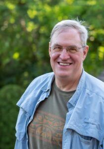 A smiling person wearing glasses, a green patterned shirt, and a light blue button-down shirt outdoors with greenery.