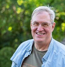 A smiling person wearing glasses, a green patterned shirt, and an unbuttoned blue overshirt, standing outdoors.