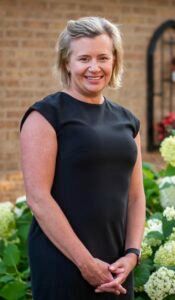 A person with short blonde hair wears a black dress, smiling with hands clasped in front of them by a brick wall and plants.