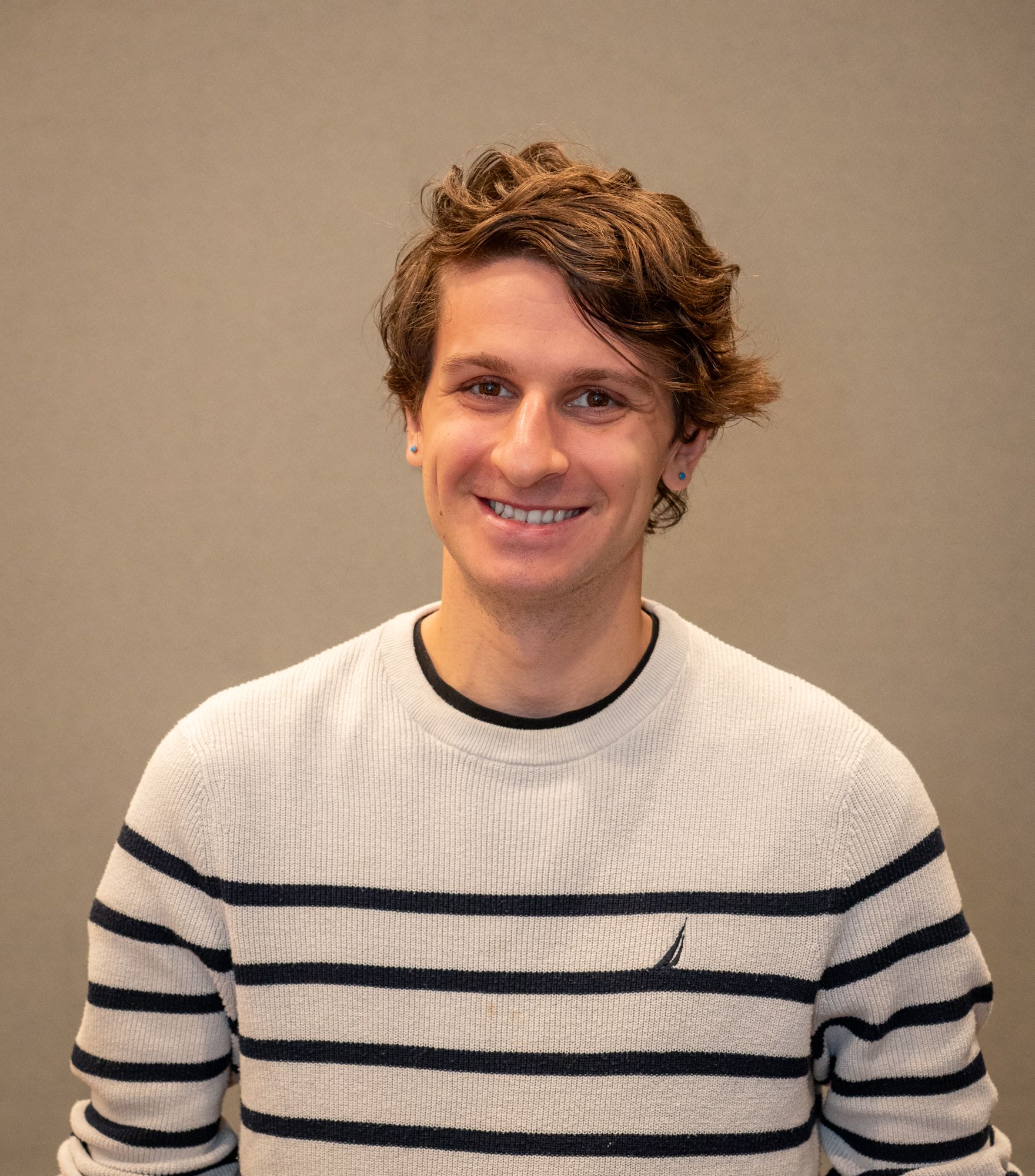 A person with brown hair smiles, wearing a white and dark blue striped crewneck sweater against a plain grey background.