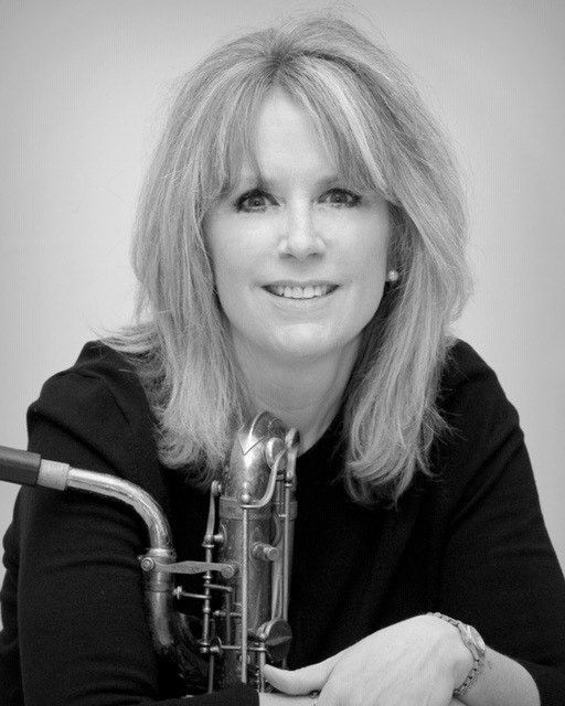 A woman smiling softly while holding a saxophone against a plain background.