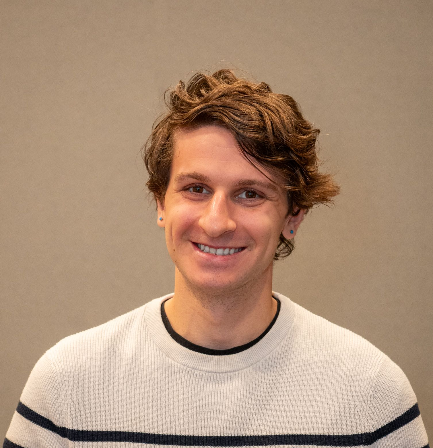A smiling person with wavy brown hair wearing a white and dark blue striped sweater against a plain gray background.