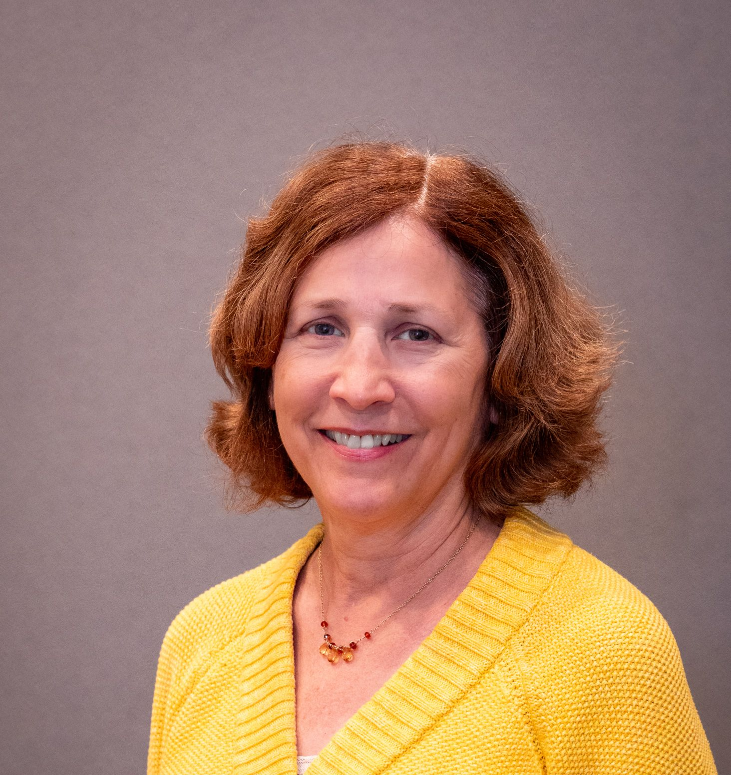 A person with shoulder-length wavy brown hair smiling, wearing a yellow knitted cardigan and a simple necklace.
