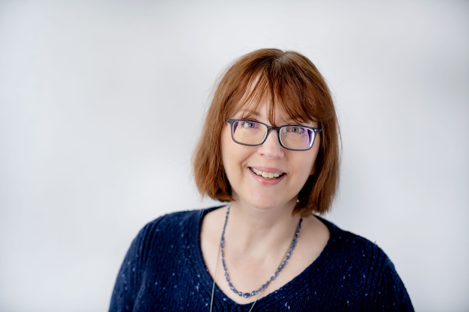 A woman with shoulder-length auburn hair and glasses smiling against a plain white background.