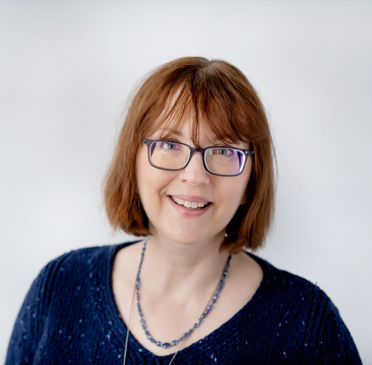 A person with shoulder-length auburn hair and glasses smiling against a white background, wearing a dark blue sweater.