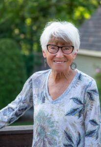 A person with short white hair and glasses wearing a floral-print top, smiling outdoors.
