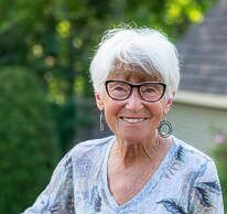 A person with short white hair and glasses smiles while wearing a long-sleeved shirt with a blue and white leafy pattern.
