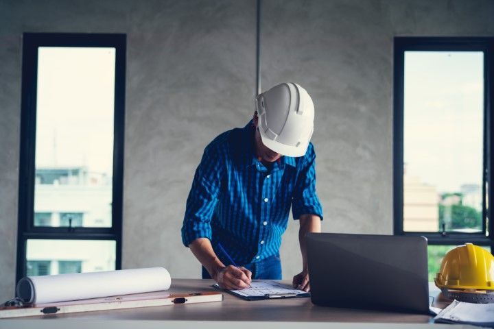 Construction worker in a blue shirt and hard hat, reviewing plans at a desk with a laptop and rolled blueprints.