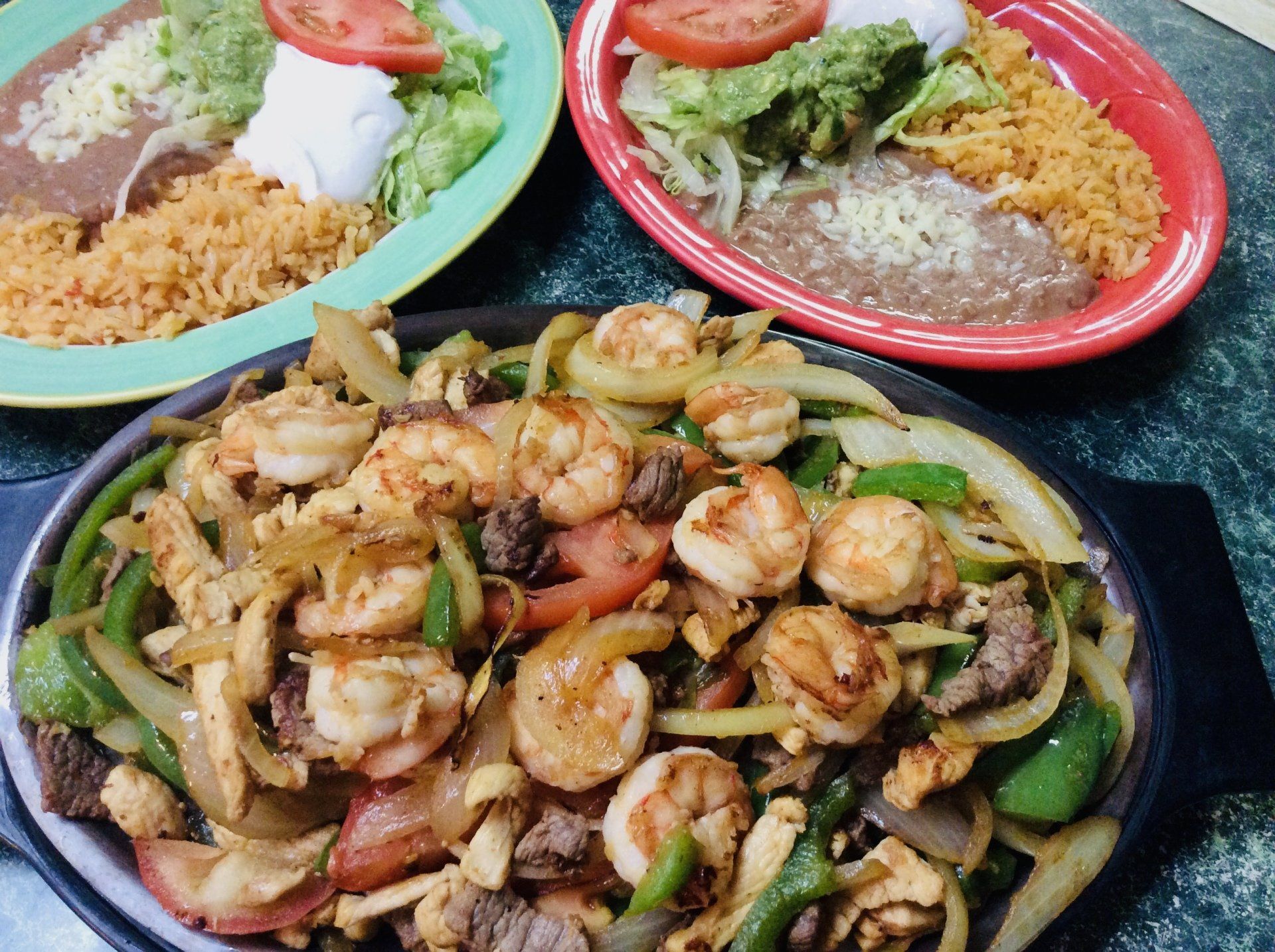 Sizzling fajita platter with shrimp, chicken, beef, vegetables. Rice and bean sides on plates.
