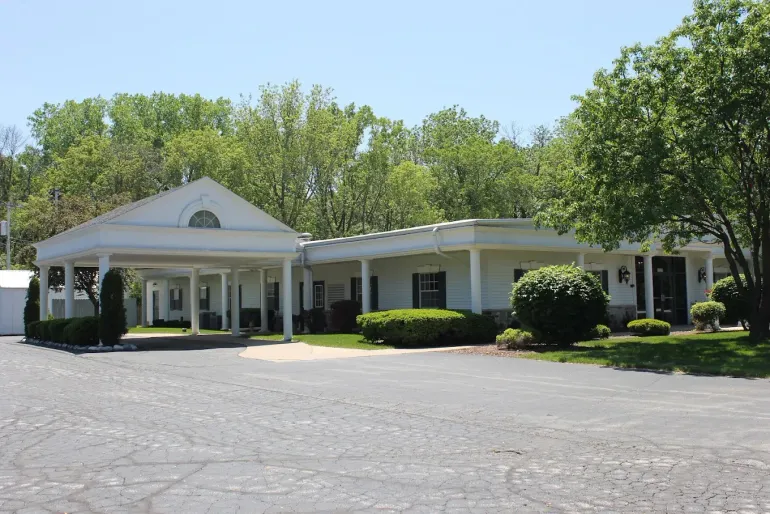 A single-story building with a carport, surrounded by trees and a paved parking area.