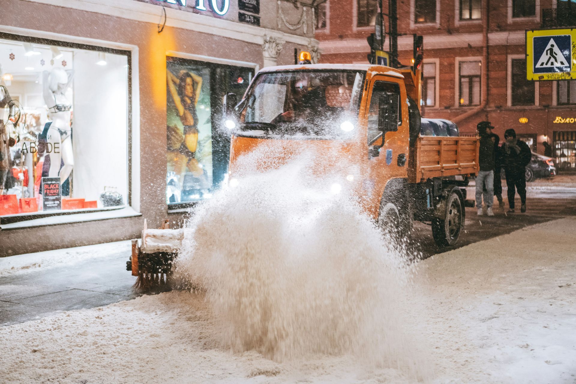 Orange snowplow clearing snow on a snowy city street at night, with pedestrians nearby