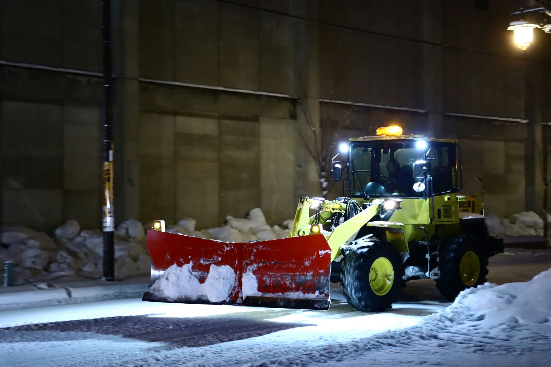Yellow snowplow clearing snow on a snowy street at night, with red blade and bright headlights