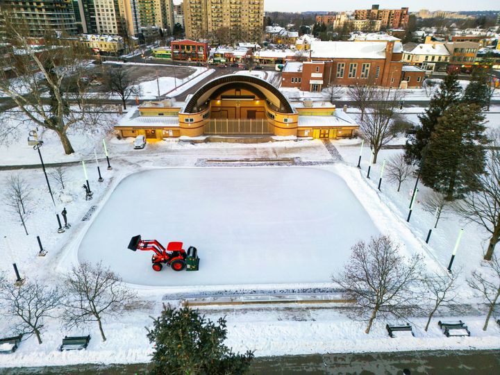 Snow-covered outdoor ice rink with a red Zamboni grooming the ice near a band shell in a park