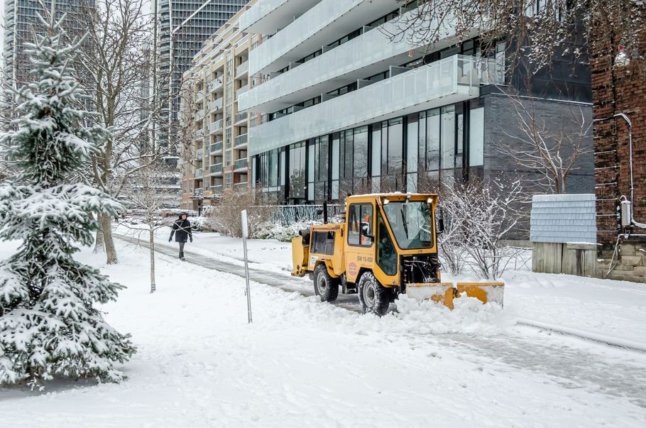 Yellow snowplow clearing a snowy sidewalk beside a modern building in winter