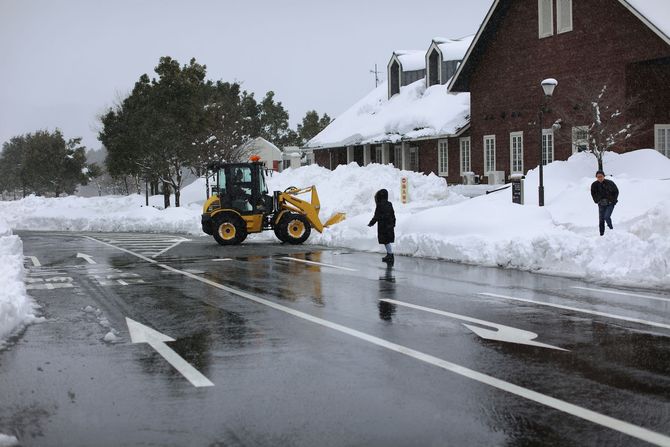 Snow-covered street with a yellow snowplow clearing slush near houses and pedestrians.