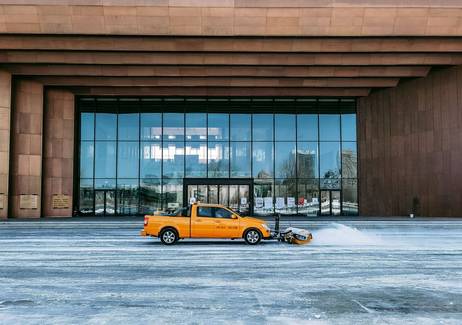 Orange snowplow truck in front of a modern building with large glass doors