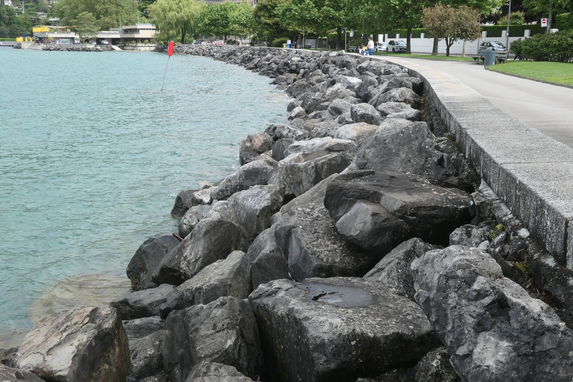 Das Ufer des Genfer Sees, rechts ein asphaltierter Weg, dann eine Steinmauer, davor hohe Aufschüttungen von großen Steinen, bevor das Wasser links kommt. 