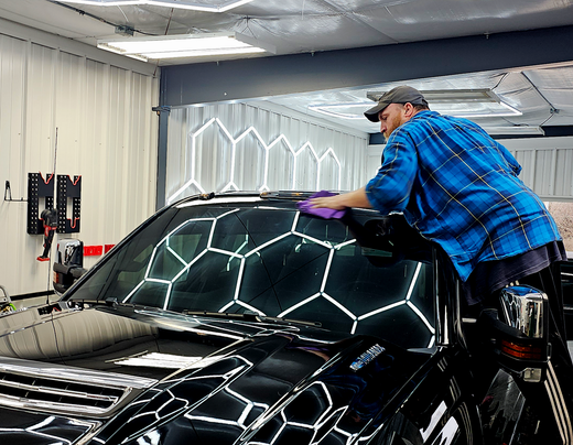 A technician wearing a blue plaid shirt stands on a step stool, applying ceramic coating to a prepped surface.