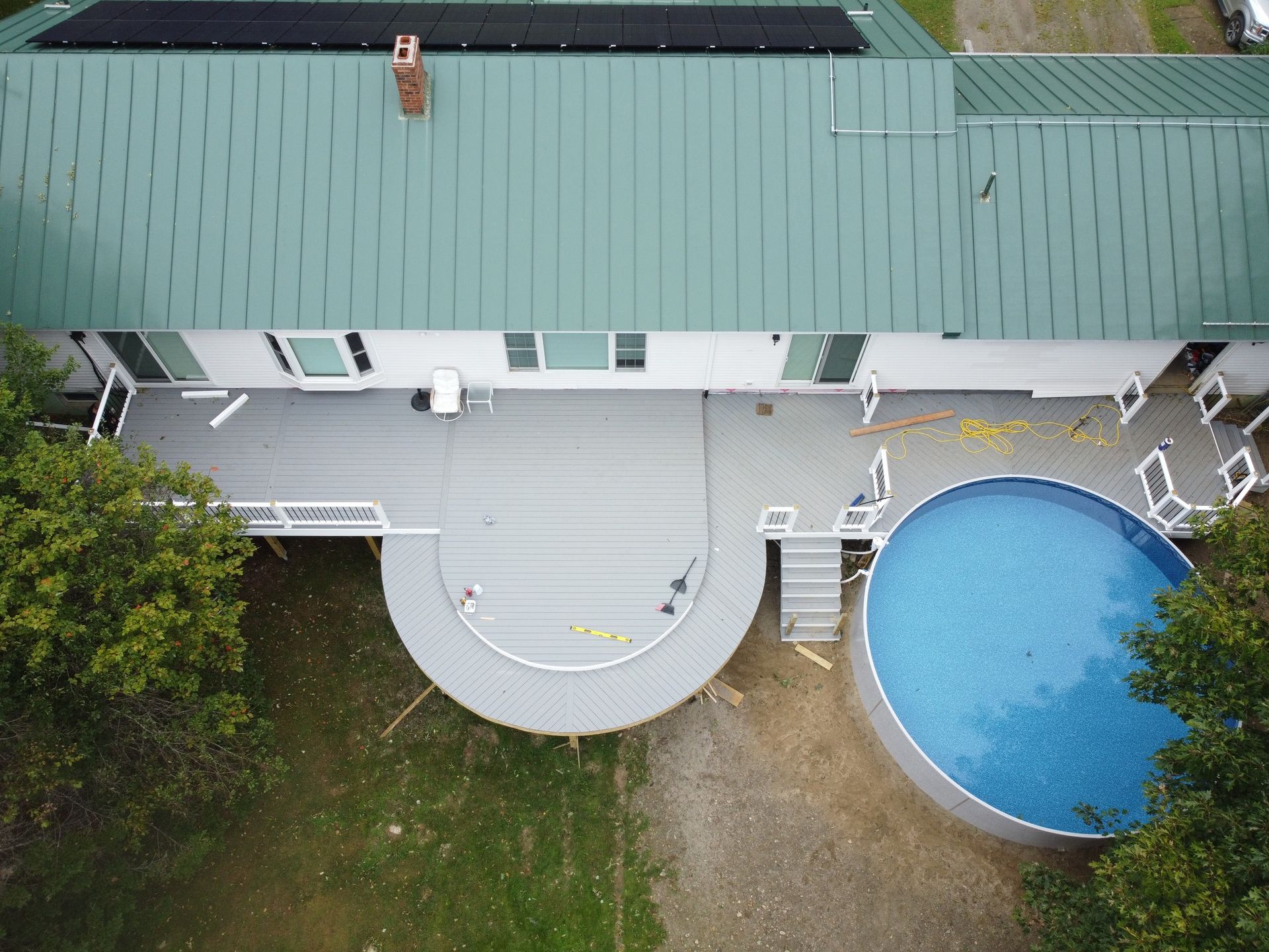 Aerial view of a house with a green roof, curved gray deck, and round blue pool beside a yard.