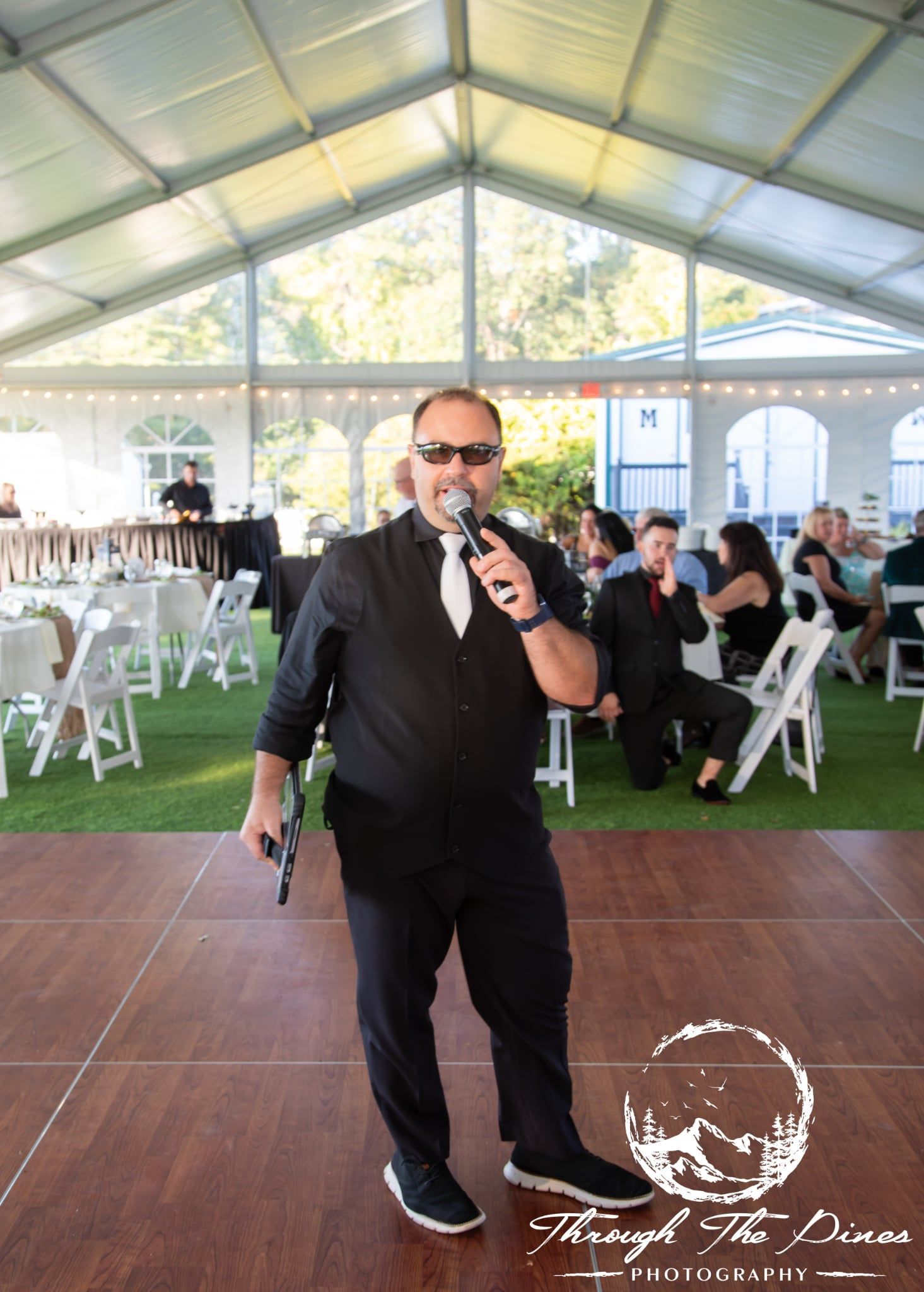 Man in black suit with microphone on dance floor, event tent in background.
