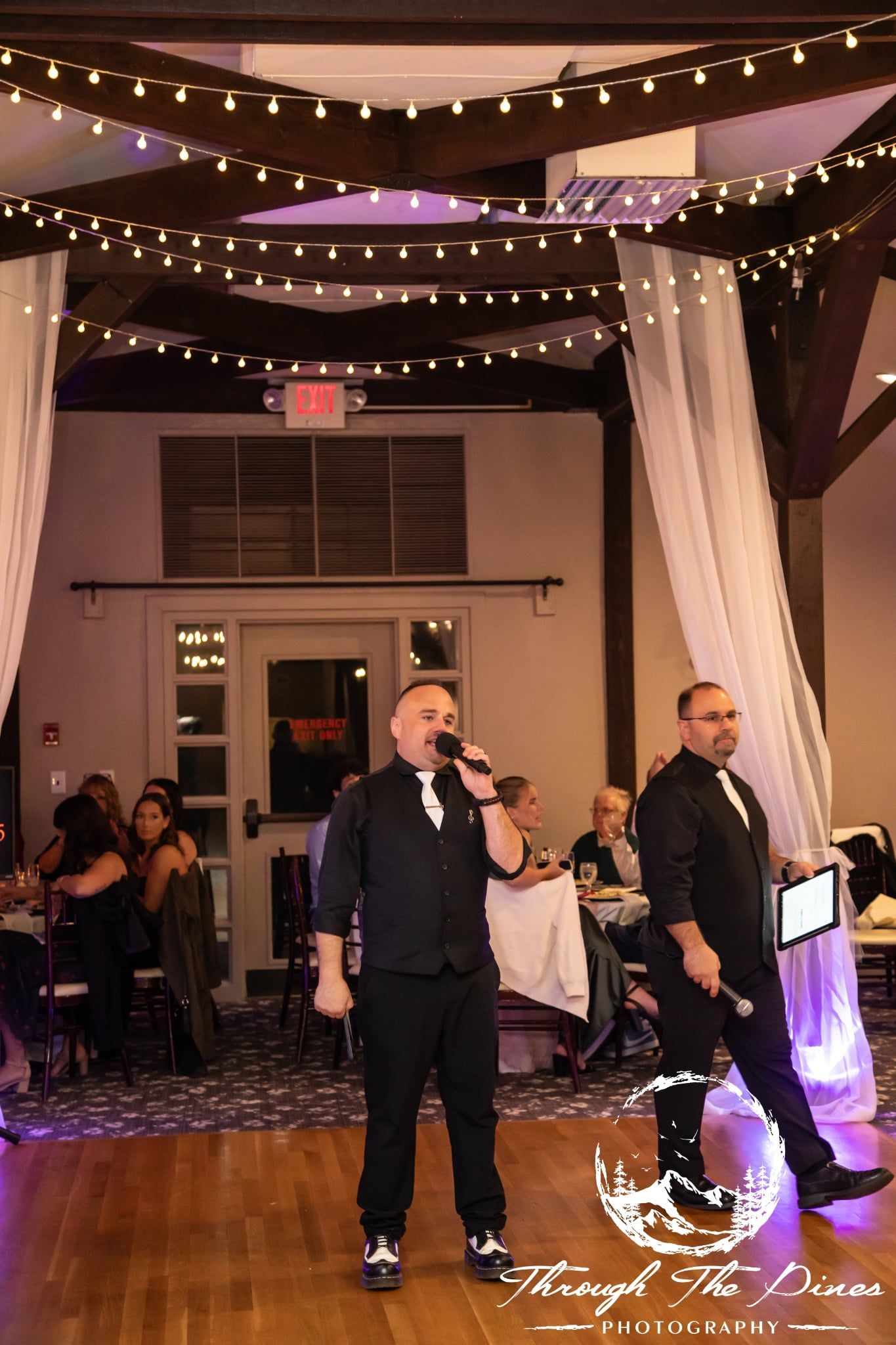 Two men in black vests singing on a stage, fairy lights overhead.