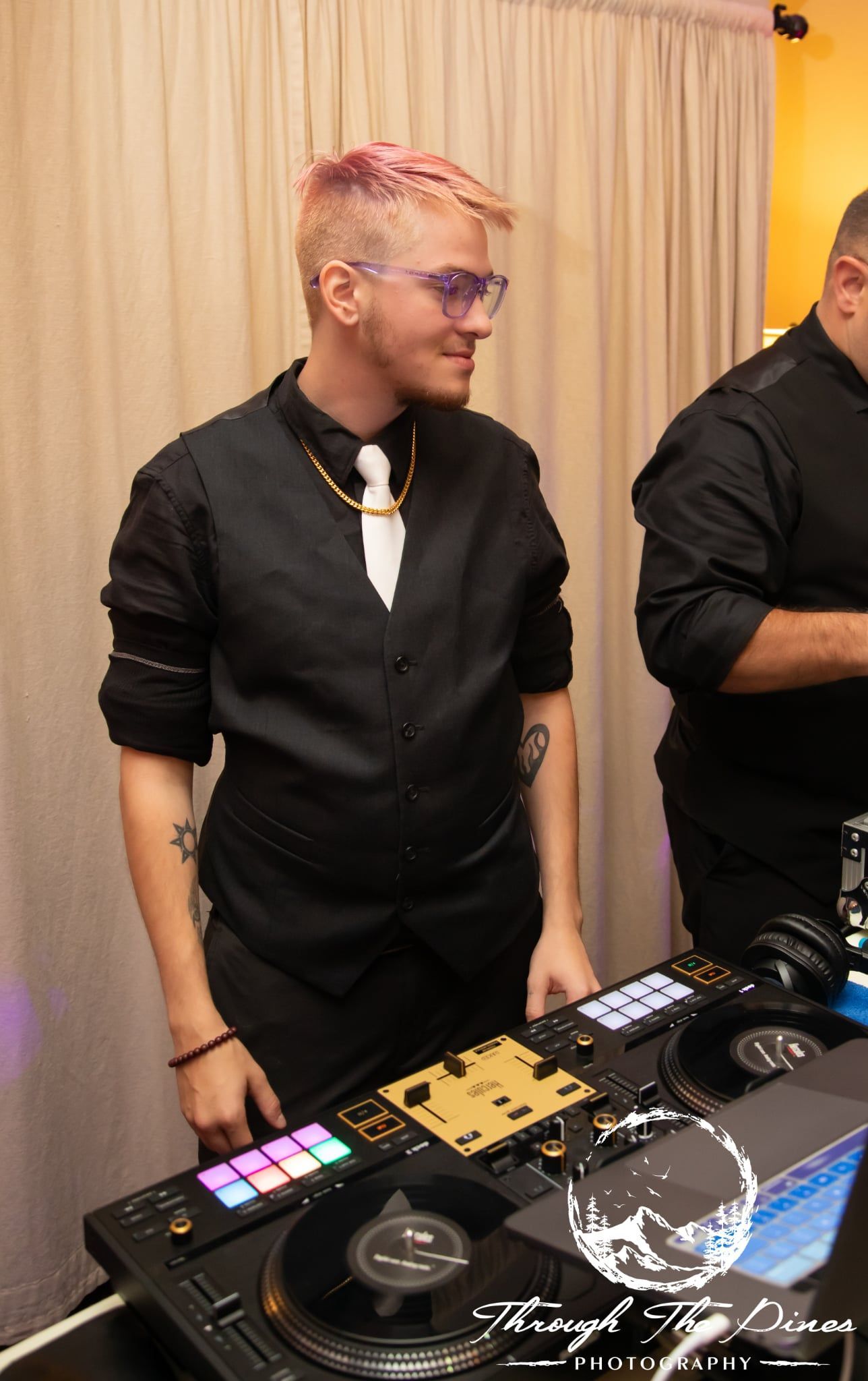 DJ with pink hair and glasses at a console, wearing a black vest and white tie.