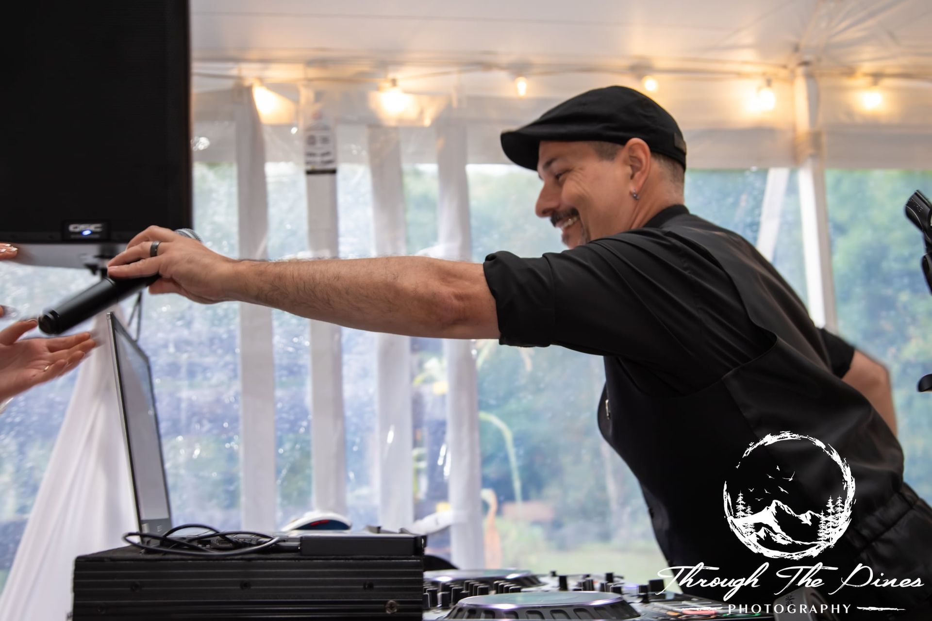 DJ in black, handing a microphone. Tent setting with a laptop and sound equipment.