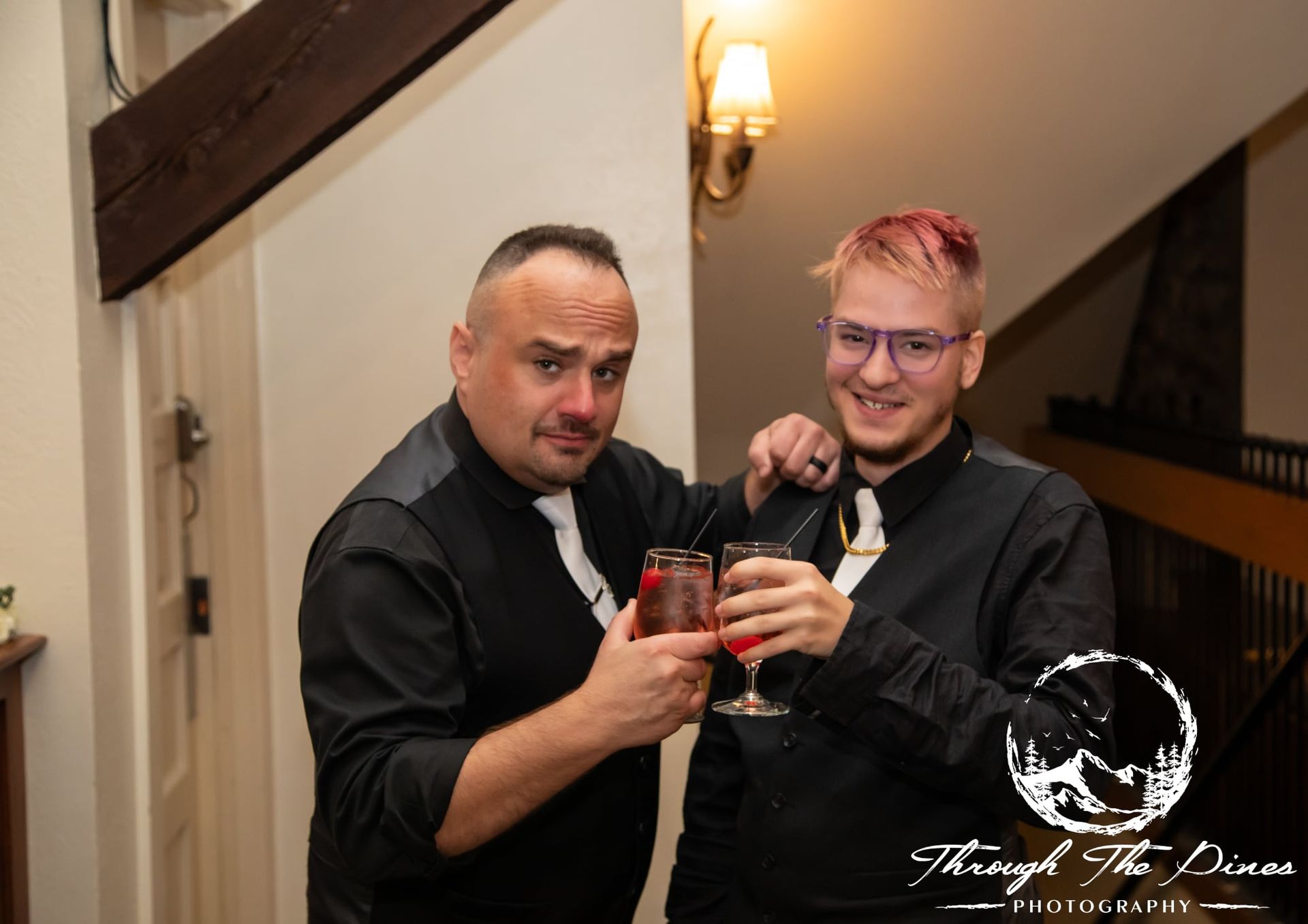 Two people in black shirts clink glasses, smiling, inside a building.