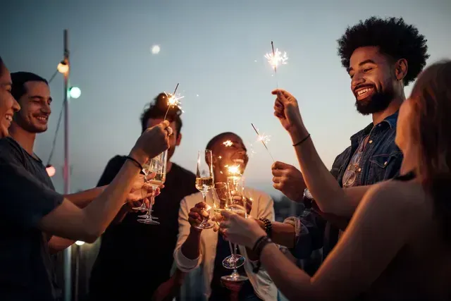 People on a rooftop celebrate, toasting champagne, holding sparklers under a dusky sky with string lights.