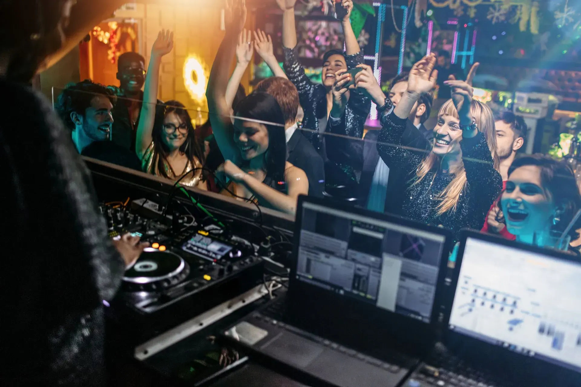 DJ at console with a crowd dancing and raising hands in a brightly lit nightclub.