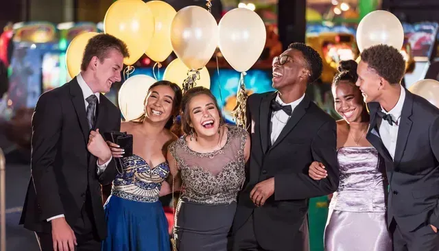 Group of formally dressed teens laughing, arm in arm, near balloons and arcade games.