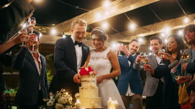Couple cutting wedding cake at outdoor reception, guests toasting. Evening setting with string lights and candles.