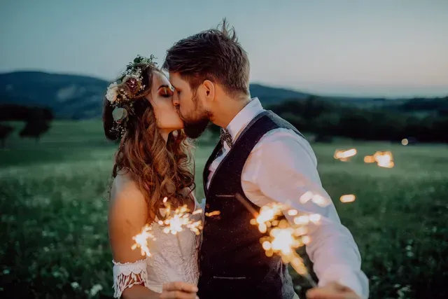 Couple kissing, holding sparklers in a field at dusk. Bride wears flower crown, groom in vest.