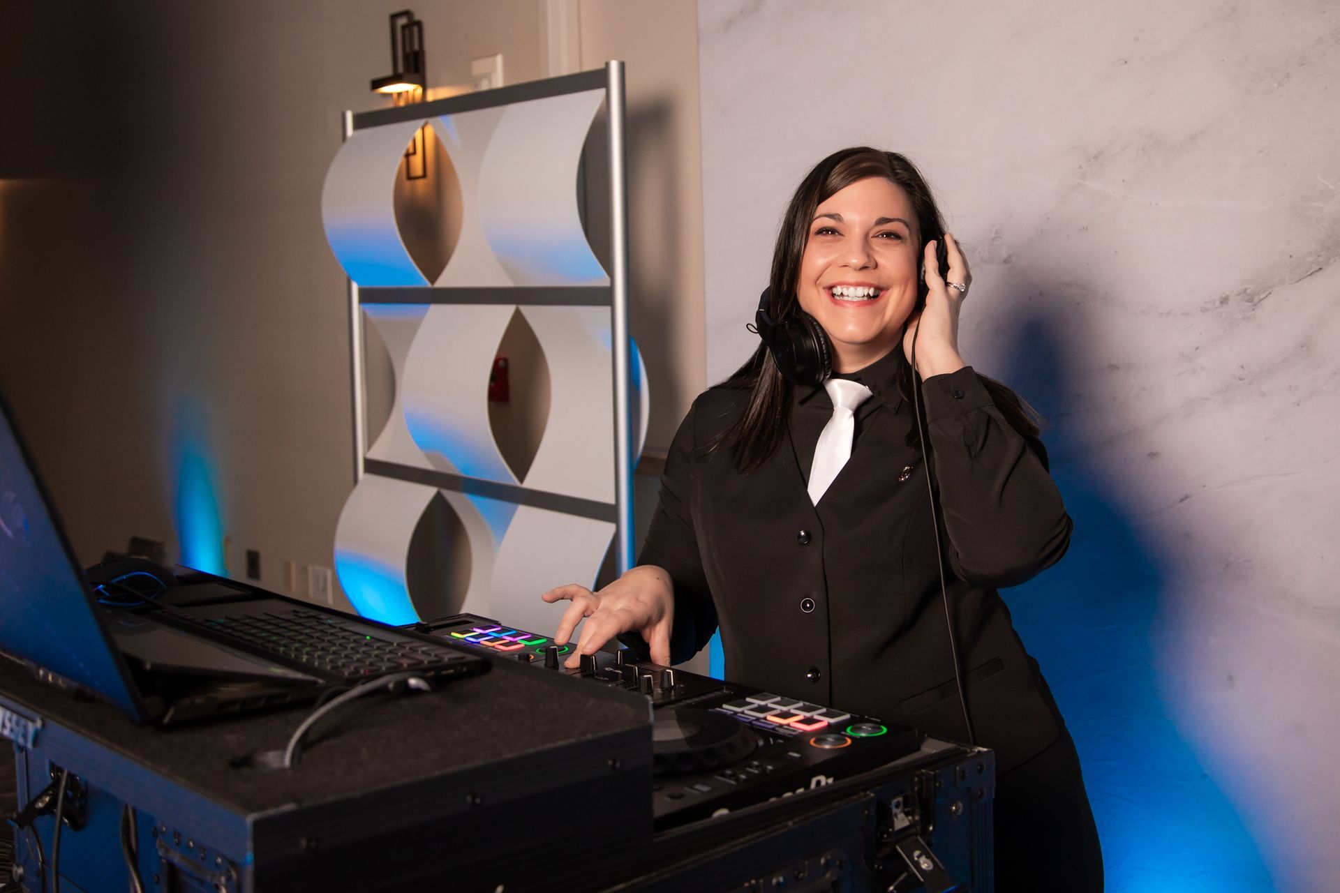 Woman in black suit smiles, adjusts headphones while DJing at a console with blue accent lighting.