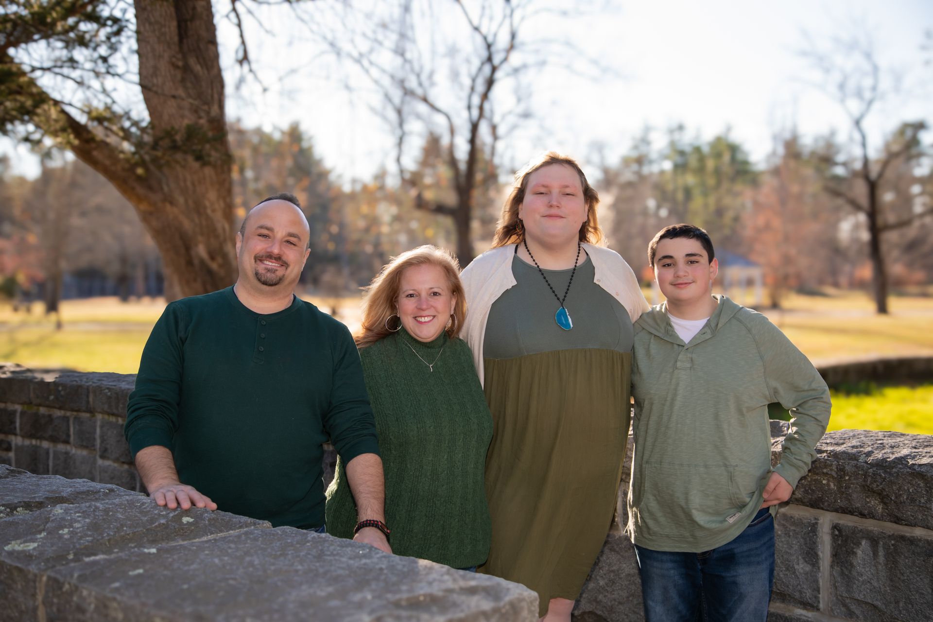 Family of four smiling outdoors, standing behind a stone wall. Green sweaters, natural background.